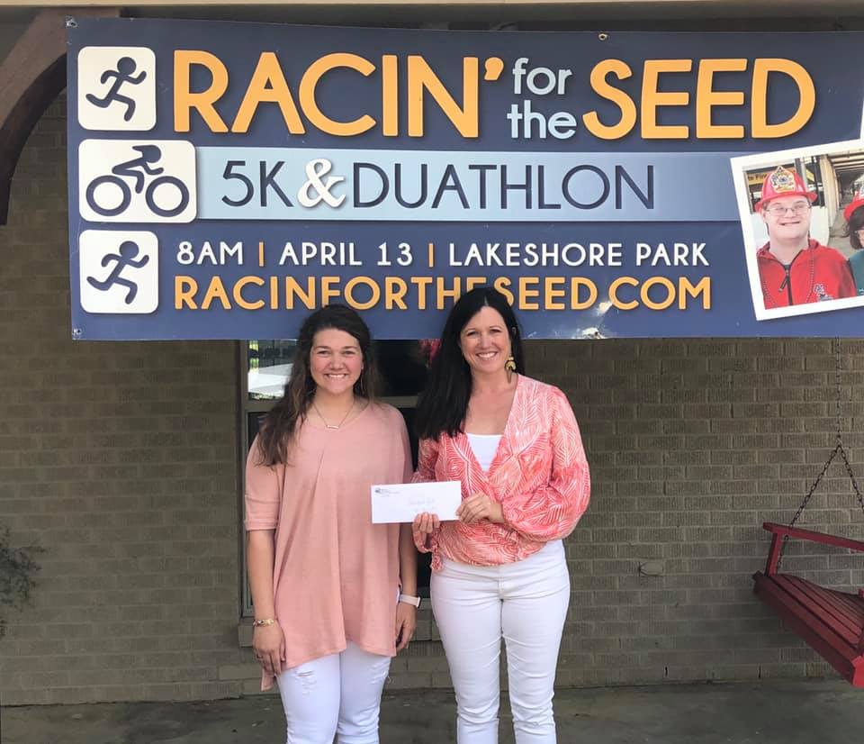 Two women pose with a check in front of the Racin for the Seed 5k and duathlon poster.